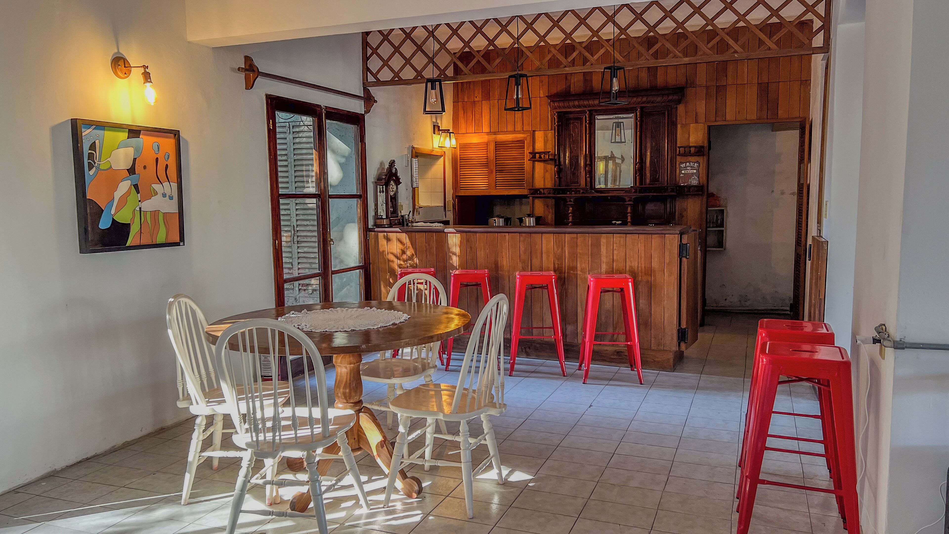 A dining area with a wooden table and white chairs, a kitchen with wooden cabinets and red stools, and a colorful painting on the wall.