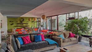 A spacious living room with a large sofa, a dining area with colorful chairs, and a view through large windows.