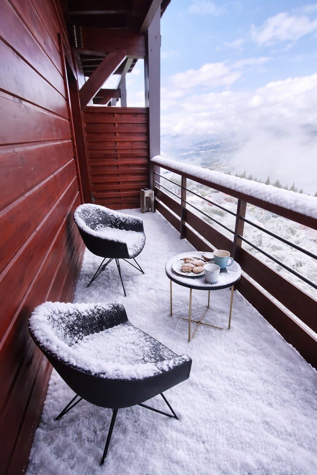Un balcón cubierto de nieve con dos sillas y una mesa pequeña, con vistas a un paisaje nevado.