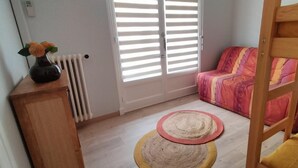 Una habitación con una cama, un radiador, una ventana con persianas, una silla y una pequeña mesa con un florero.