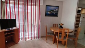Una sala de estar con televisor sobre un mueble de madera, una mesa de comedor con sillas, y un área de cocina con armarios y electrodomésticos.