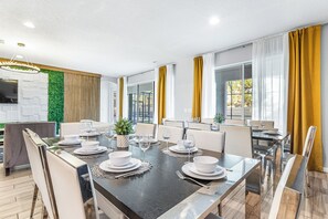 A modern dining area with a long table set for six, surrounded by chairs. - UpstaySpaciousHomePoolHotTubSleeps27 (Kissimmee)