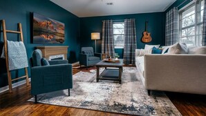 A living room with a dark blue wall, a fireplace, a ceiling fan, a guitar, and a large window with curtains. - The SpokeEasy DownTown Inn (Bentonville)