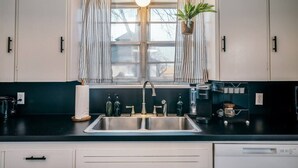 A kitchen with a double sink, a hanging light fixture, a window with curtains, and a potted plant. - The SpokeEasy DownTown Inn (Bentonville)