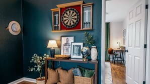 A cozy room with a dartboard, framed pictures, a lamp, and a console table with cushions and a globe. - The SpokeEasy DownTown Inn (Bentonville)