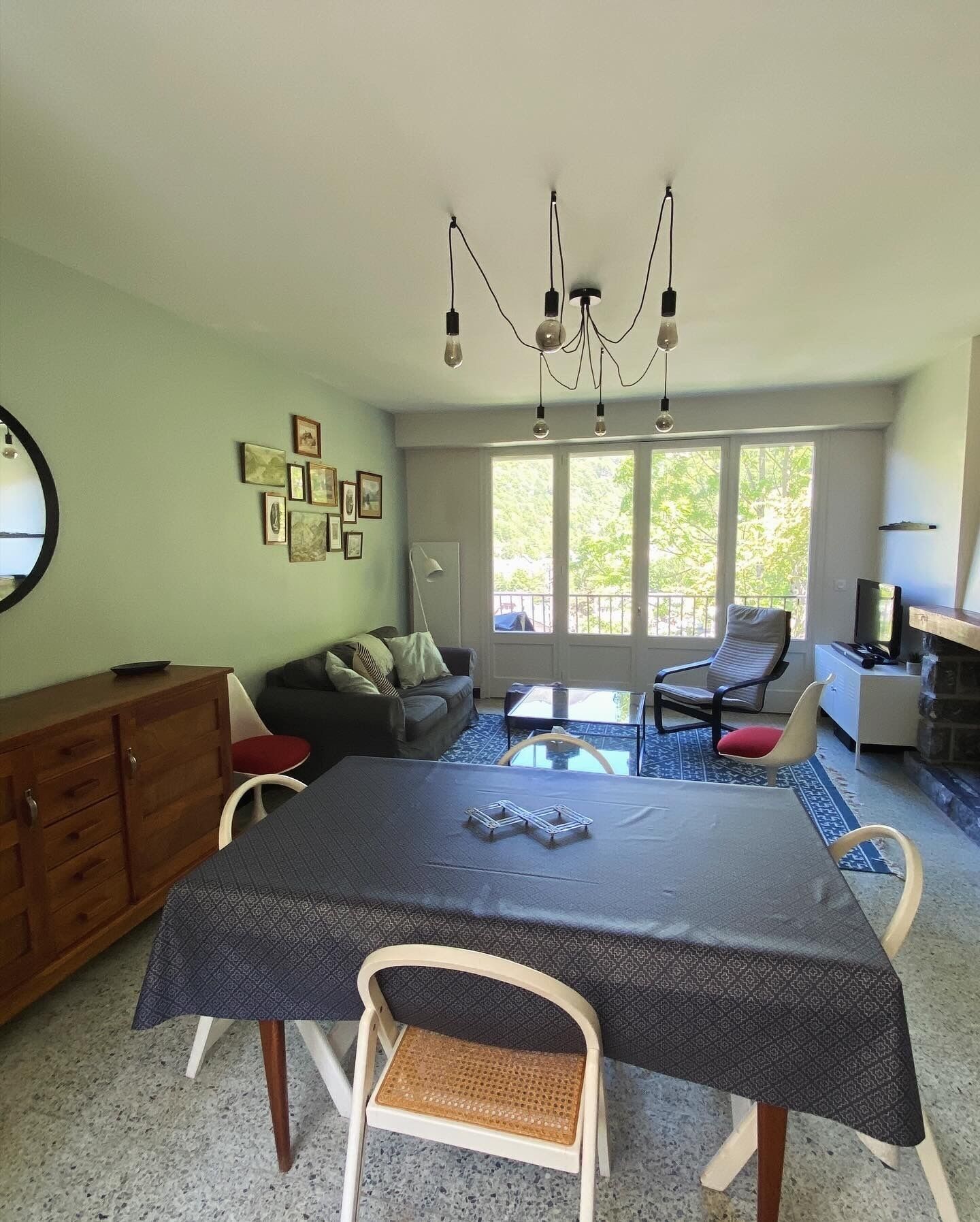 A dining area with a table, chairs, a chandelier, and a view through large windows.