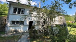 Une maison à deux étages avec une façade en pierre, une cheminée bien visible et un petit jardin agrémenté d’un sentier menant à l’entrée.