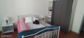 A neatly made bed with a patterned bedspread and pillows, a bedside table with a lamp, and a wardrobe with a curtain. - Maison Proche D'avignon, L'isle /sorgues, Saint Remy et du Parc Spirou (Cavaillon)