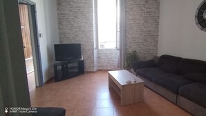 Un soggiorno con un divano scuro, un tavolino in legno, una TV a schermo piatto su un mobile e una sveglia a parete.