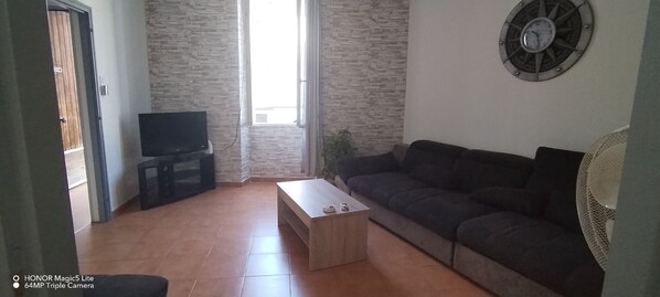 A living room with a dark-colored sofa, a wooden coffee table, a flat-screen TV on a stand, and a wall clock. - Maison Proche D'avignon, L'isle /sorgues, Saint Remy et du Parc Spirou (Cavaillon)
