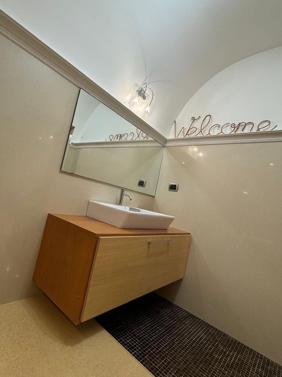 A modern bathroom with a large mirror, a sink, and a 'Welcome' sign on the wall.