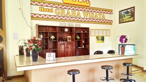 A reception desk with a sign reading "Hotel Graha Wisata" and a clock above it. There are three stools in front of the desk, and a wooden cabinet with shelves behind it. - Graha Wisata (Kotabumi)