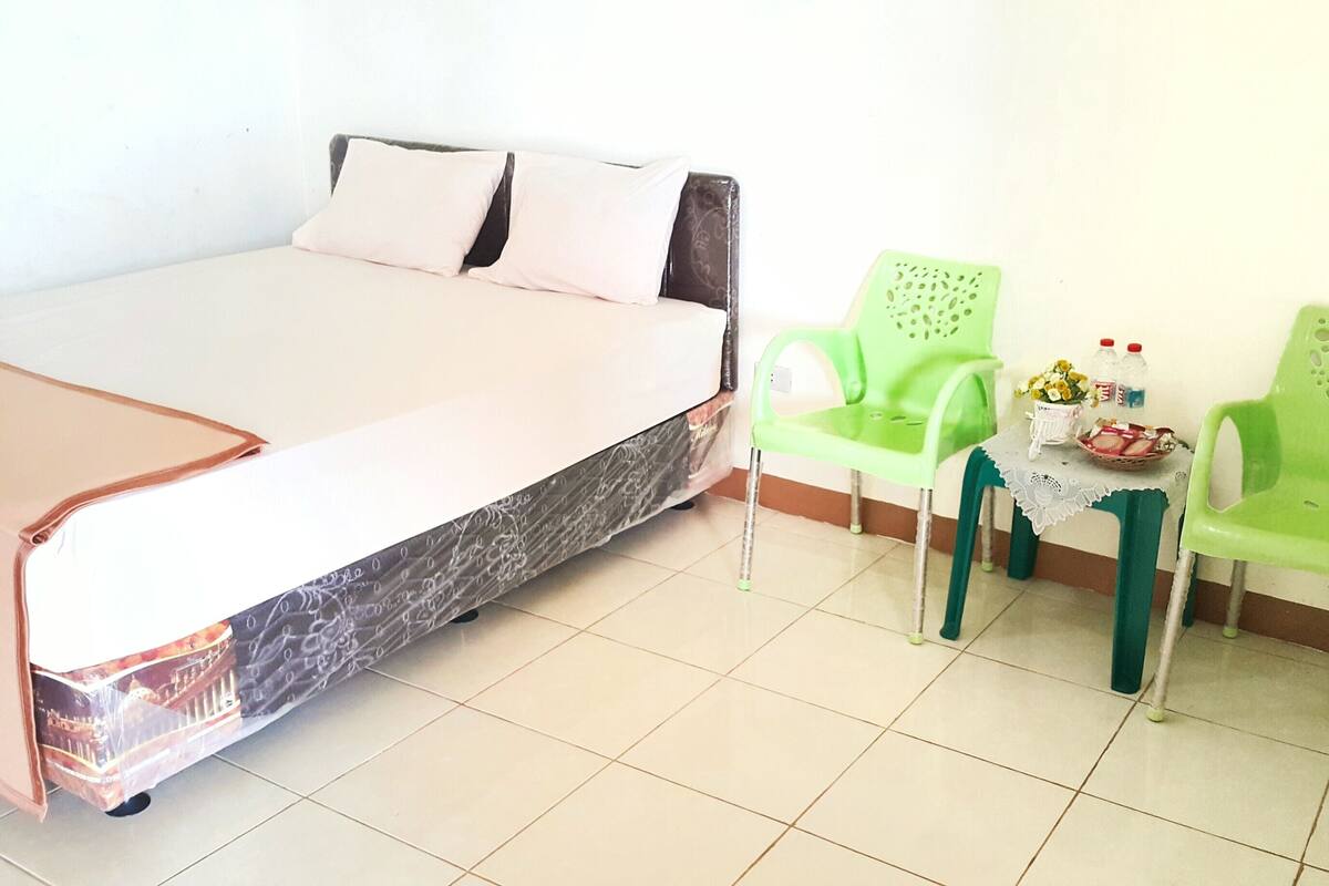A room with a bed, two green chairs, a small table with a vase and bottles, and a tiled floor.