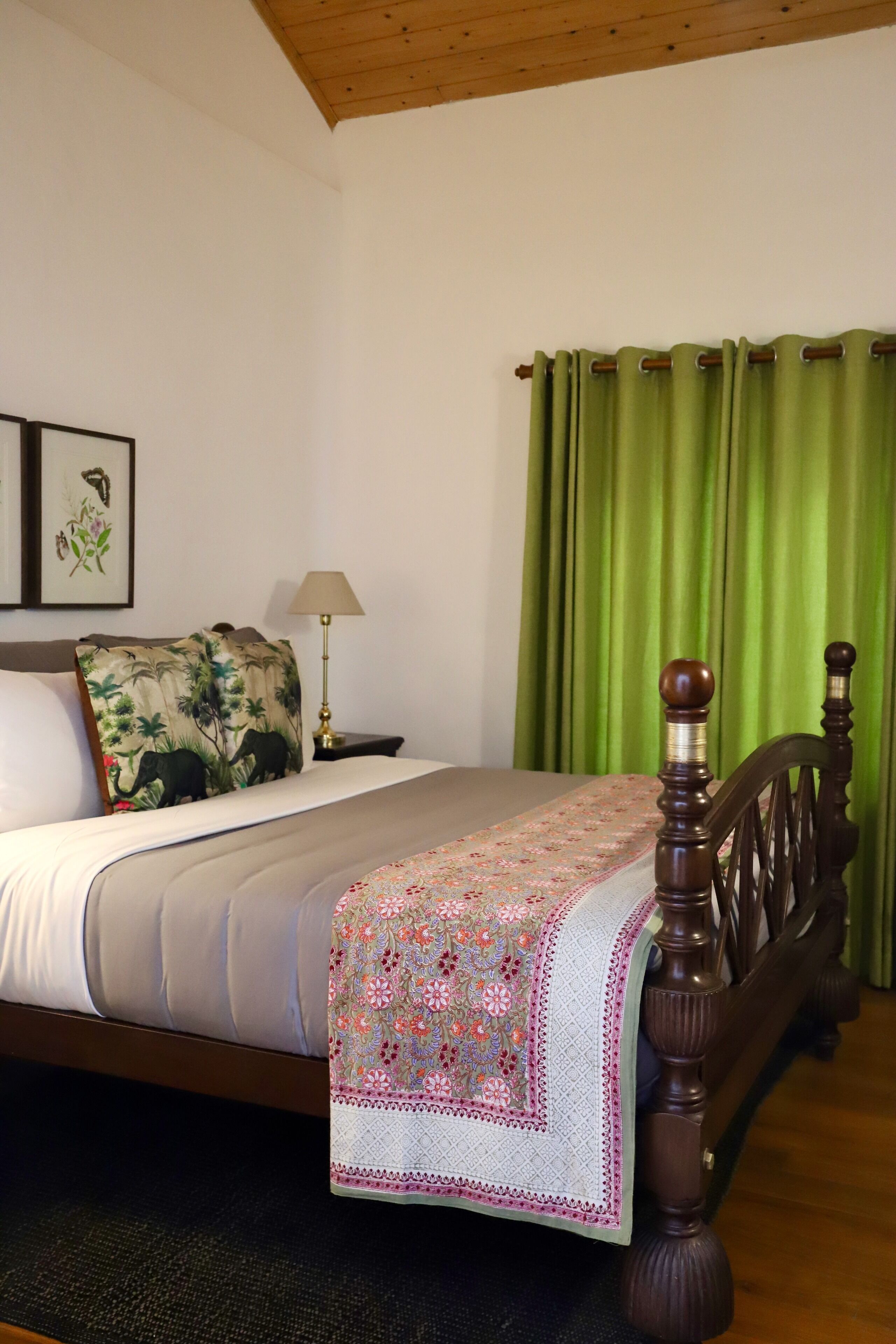 A bedroom with a wooden bed, green curtains, a nightstand with a lamp, and a wall with framed artwork.