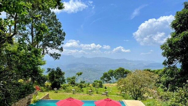 Une piscine bordée de parasols rouges, entourée d’une végétation luxuriante et offrant une vue sur des montagnes lointaines.