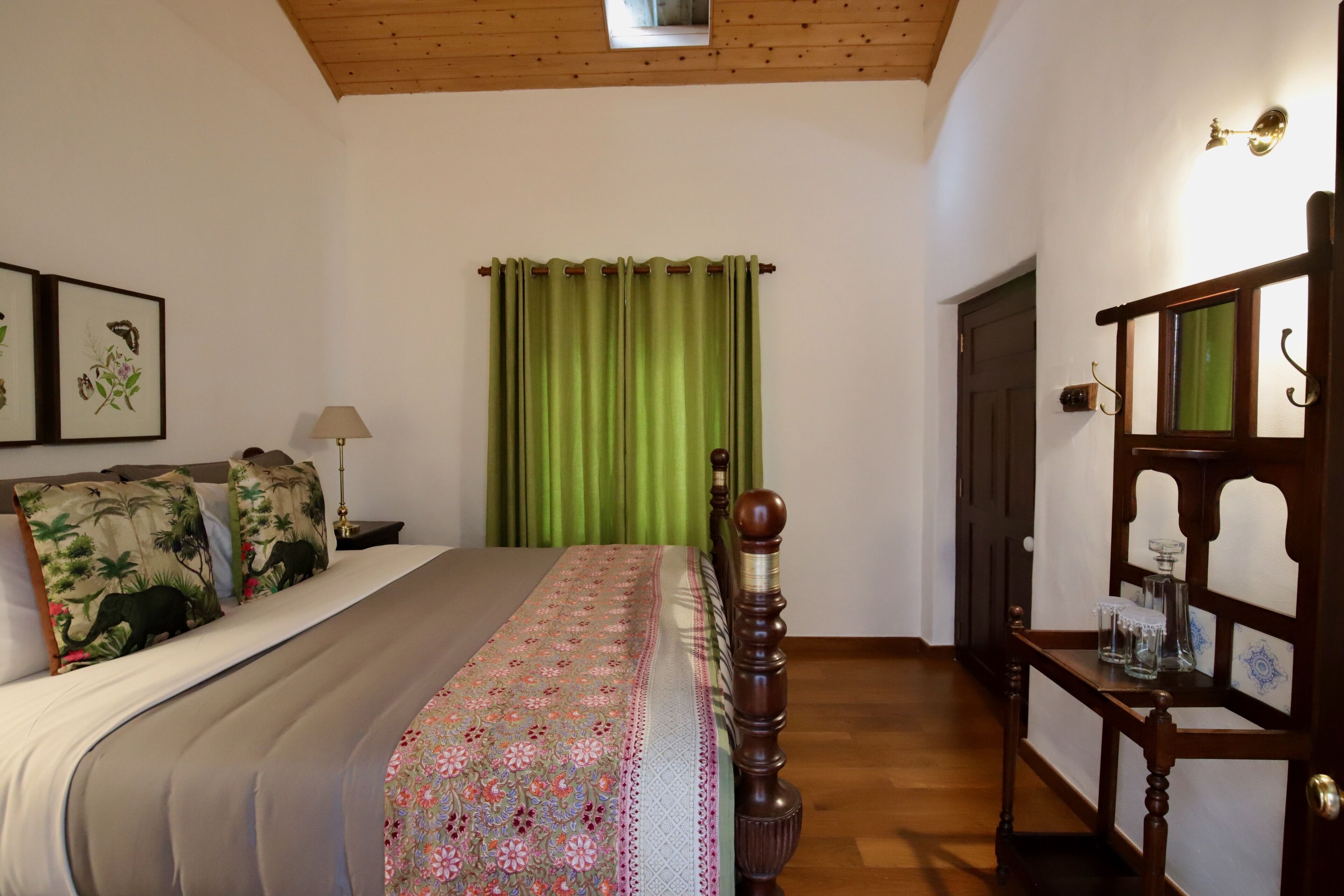 A bedroom with a bed, green curtains, a wooden headboard, a nightstand, a lamp, a dresser, and framed botanical prints on the wall.