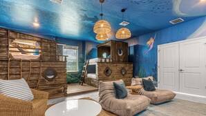 A room with a blue ceiling, wooden walls, and a mural of a mermaid. There are two brown sofas, a white table, and wicker furniture. - Upstay24-PPLHomeThemeRoomsPoolSpaCinema (Kissimmee)
