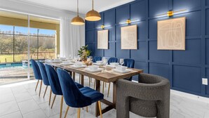 A dining area with a long table set for six, blue chairs, and a gray chair. The room has a blue paneled wall and a large window overlooking a pool. - Upstay24-PPLHomeThemeRoomsPoolSpaCinema (Kissimmee)