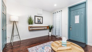 A hallway with a wooden floor, a patterned rug, a wall-mounted shelf with plants, a framed artwork, and a blue door with a window. - Cheerful home by Millwork District/Farmers Market (Dubuque)