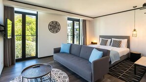 Une chambre d’hôtel moderne avec un grand lit, un canapé gris, une petite table ronde et une vue sur la verdure par les fenêtres.