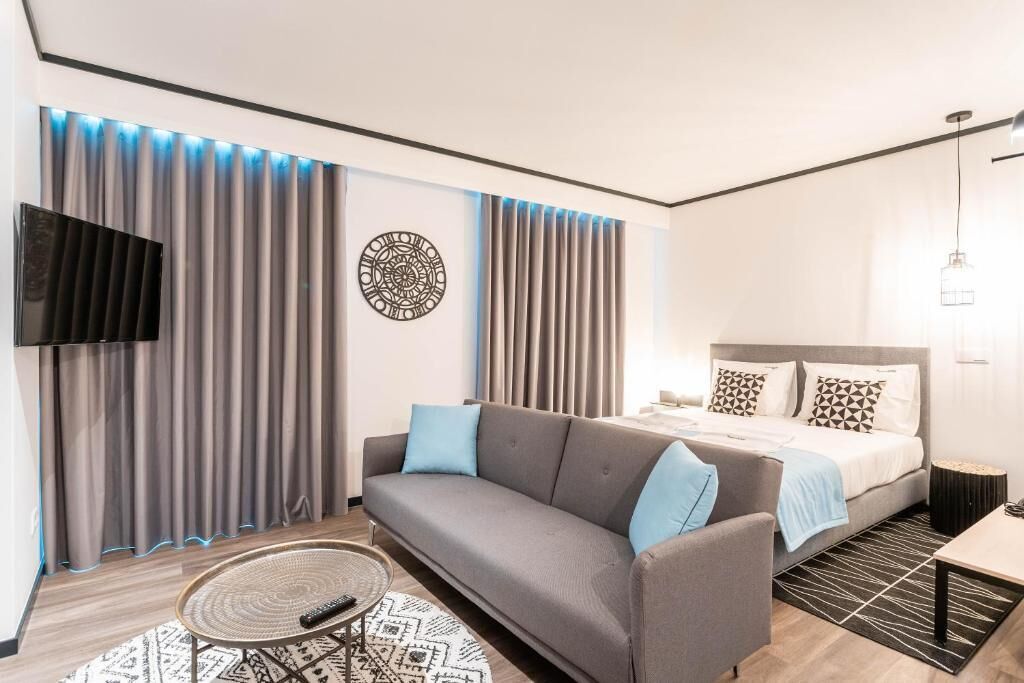 A modern hotel room with a grey sofa, a bed with patterned pillows, a flat-screen TV, and a decorative wall piece.