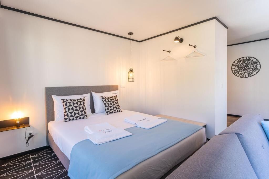 A modern hotel room with a neatly made bed, a bedside table with a lamp, a hanging light fixture, and a wall decoration.