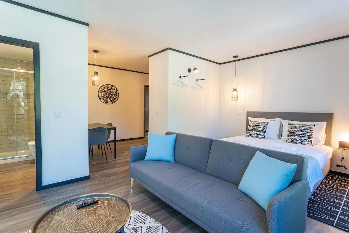 A modern hotel room with a grey sofa, a bed with patterned pillows, a dining area with a table and chairs, and a wall with a circular artwork.