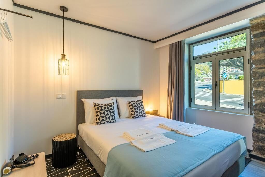 A hotel room with a bed, a bedside table, a lamp, and a window with a view of the outside.