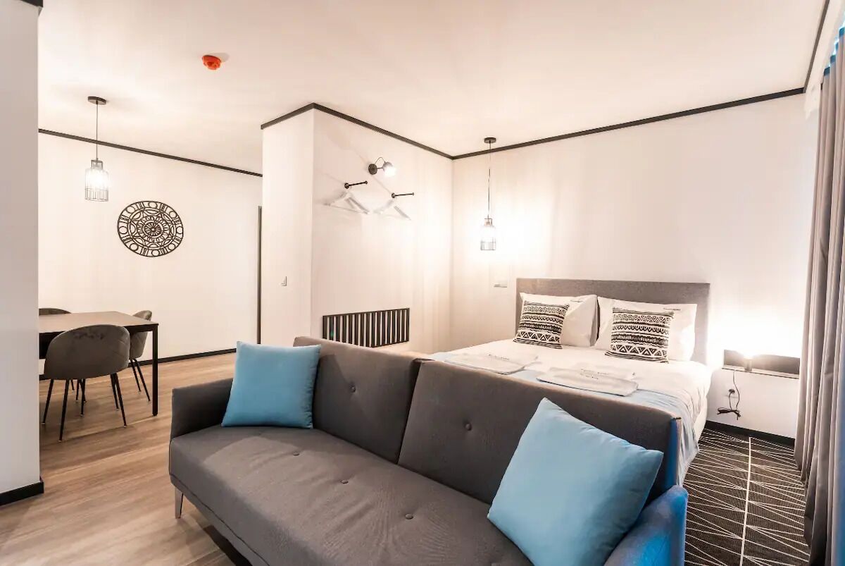 Una habitación de hotel moderna con un sofá gris, una cama con almohadones estampados, una zona de comedor con mesa y sillas, y una decoración de pared original.