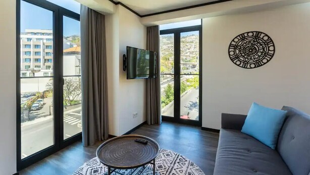 Un salon moderne doté d’une grande fenêtre, d’un téléviseur à écran plat, d’une table basse à motifs et d’un canapé gris agrémenté d’un coussin bleu.