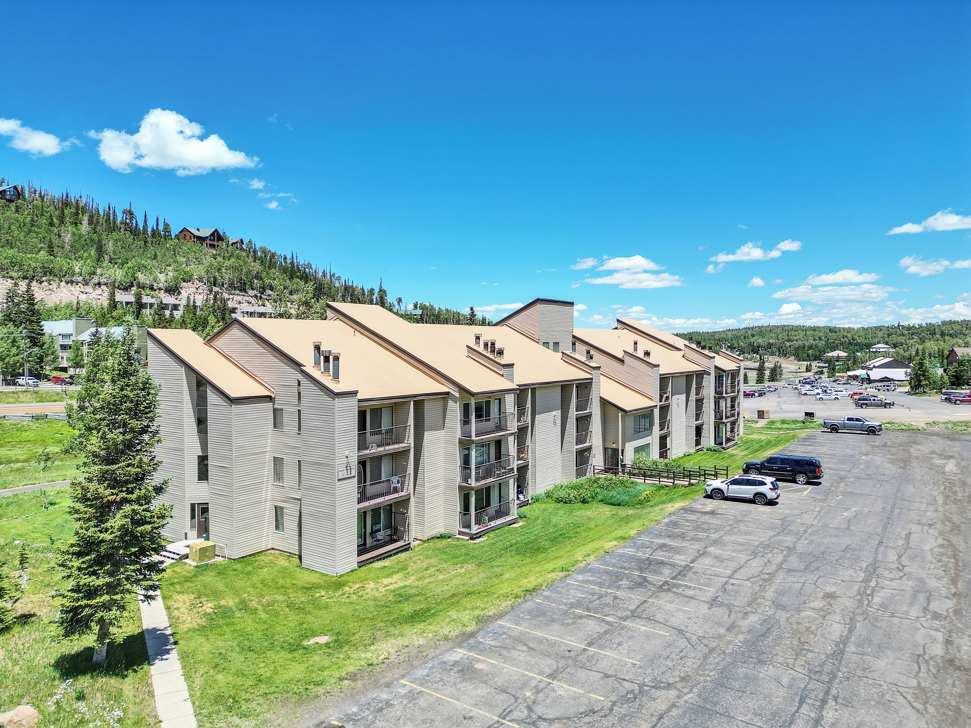 A row of multi-story buildings with balconies, a parking lot, and a green hillside in the background.