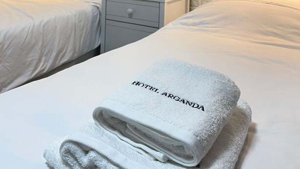 Hotelkamer met twee bedden, een nachtkastje, een ingelijste foto aan de muur en een gevouwen handdoek met 'HOTEL ARGANDA' erop.