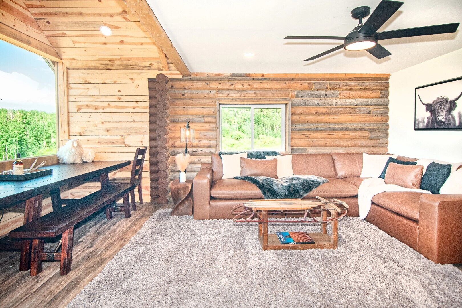 A rustic cabin interior with a wooden dining table, leather sofa, and a ceiling fan.
