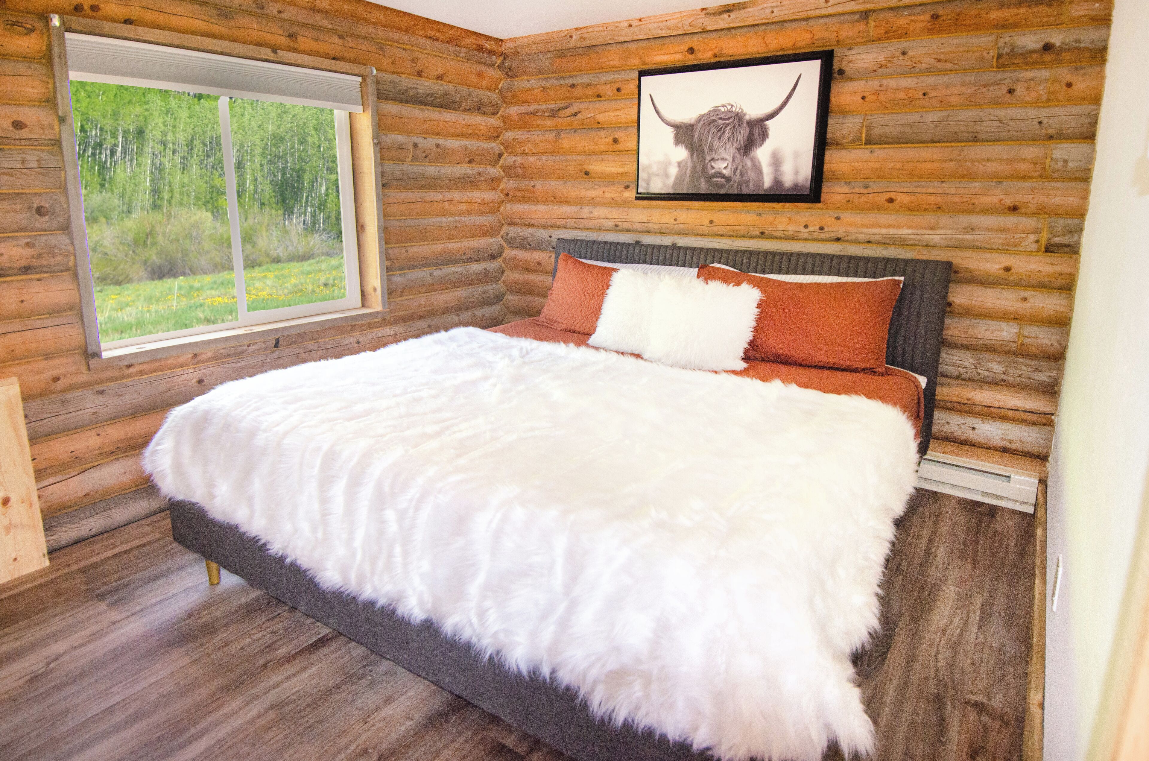 A log cabin bedroom with a large bed, a window with a view of greenery, and a framed picture of a bull on the wall.