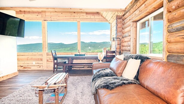 A log cabin interior with a wooden dining table, leather sofa, and a view of green hills through large windows. - EML 3 Colorow (Paonia)