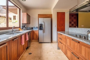 A modern kitchen with wooden cabinets, a stainless steel refrigerator, a granite countertop, and a window providing a view of the outside. - Curamoria Collection Celeste Huatulco (Santa María Huatulco)