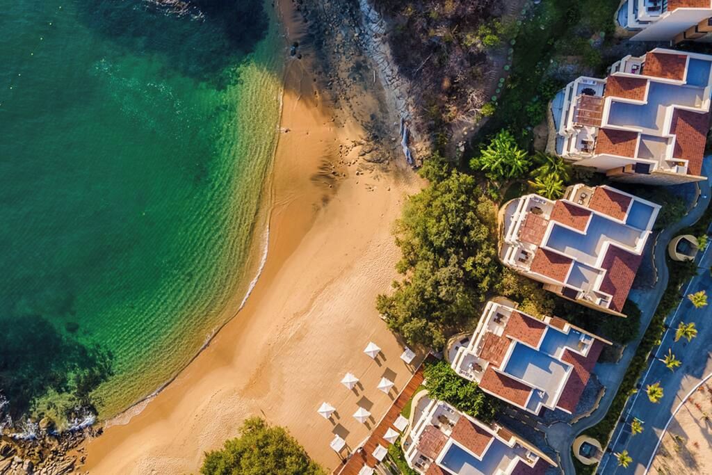 Vista aerea di una proprietà costiera con spiaggia sabbiosa, acque blu limpide e edifici residenziali con tetti in tegole rosse.