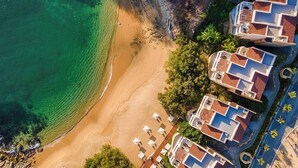 Vista aérea de una zona costera con una playa de arena, un mar azul cristalino y edificios con techos de tejas rojas.