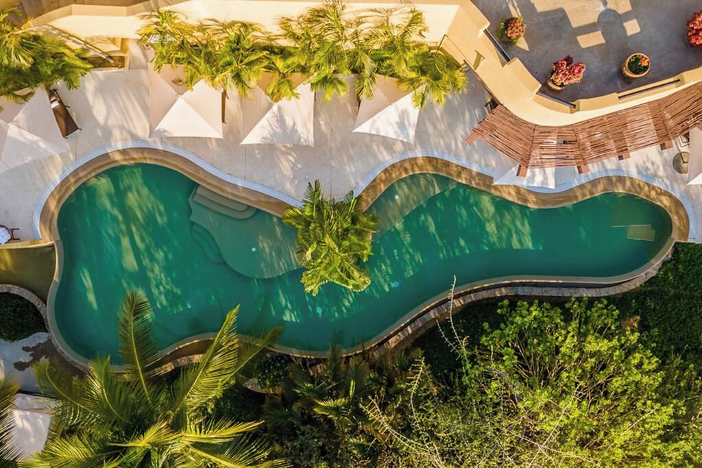A large, uniquely shaped swimming pool surrounded by lush greenery and white structures.