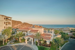 A coastal residential area with multiple buildings, including a prominent white structure with a red-tiled roof, surrounded by palm trees and a clear view of the ocean.