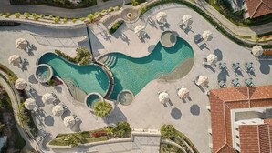Ovenudblik over en ferieanlæg med en stor central pool, flere loungeområder med parasoller og et velplejet landskab.