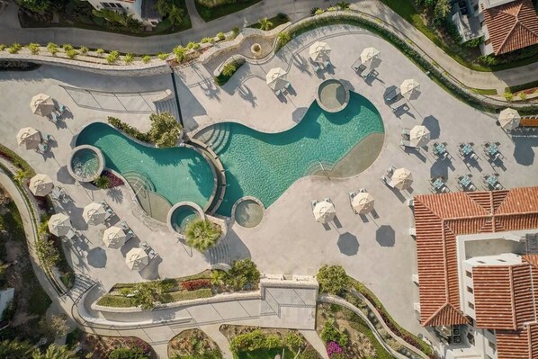 An aerial view of a resort with a large central pool, multiple lounge areas with umbrellas, and a well-maintained landscape.