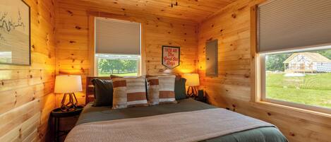 A wooden cabin bedroom with a bed, two bedside lamps, a ceiling fan, and two windows with blinds.