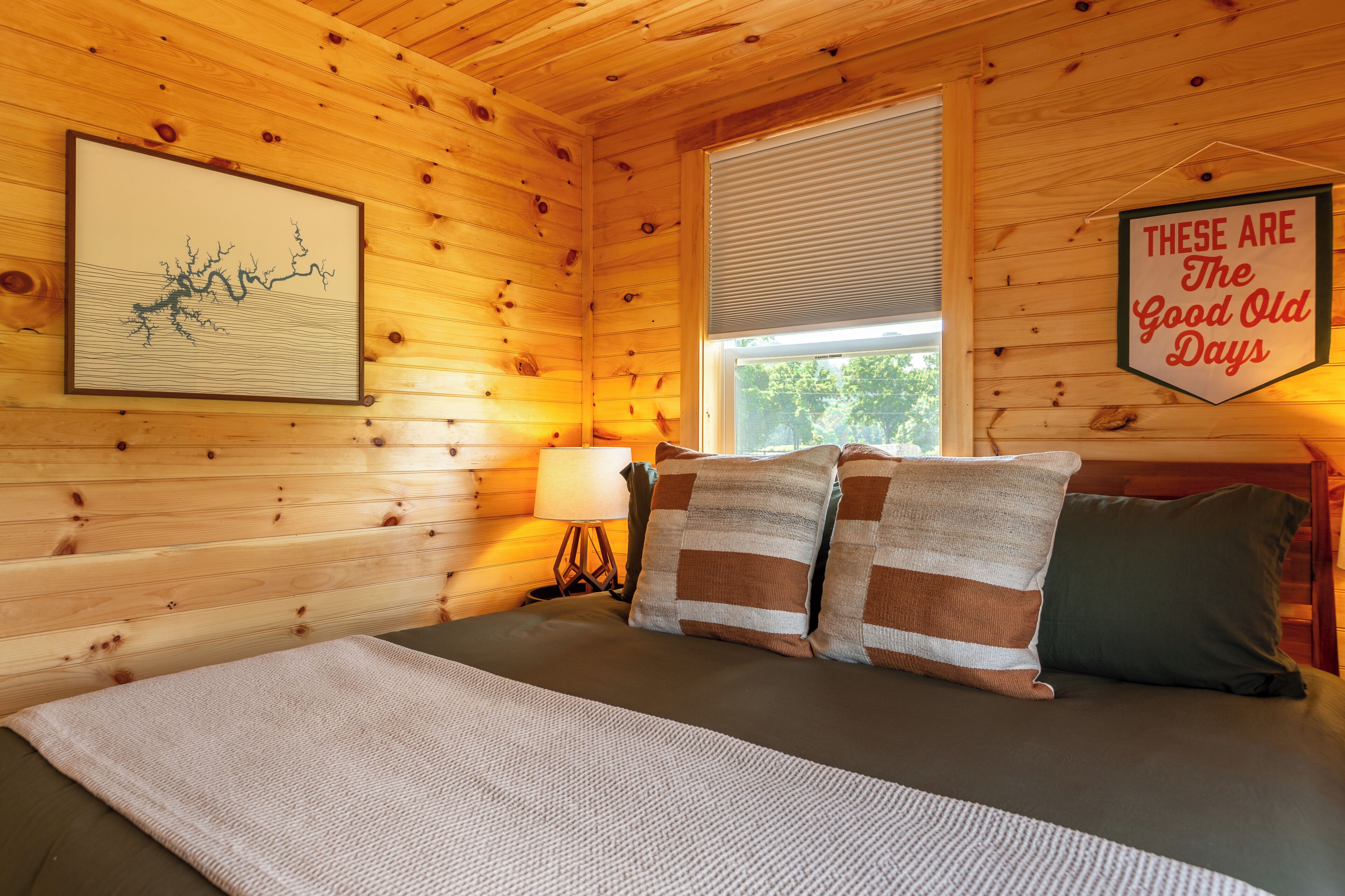 Una habitación de cabaña de madera con una cama, una obra de arte enmarcada, un ventanal con persianas y un cartel en la pared.