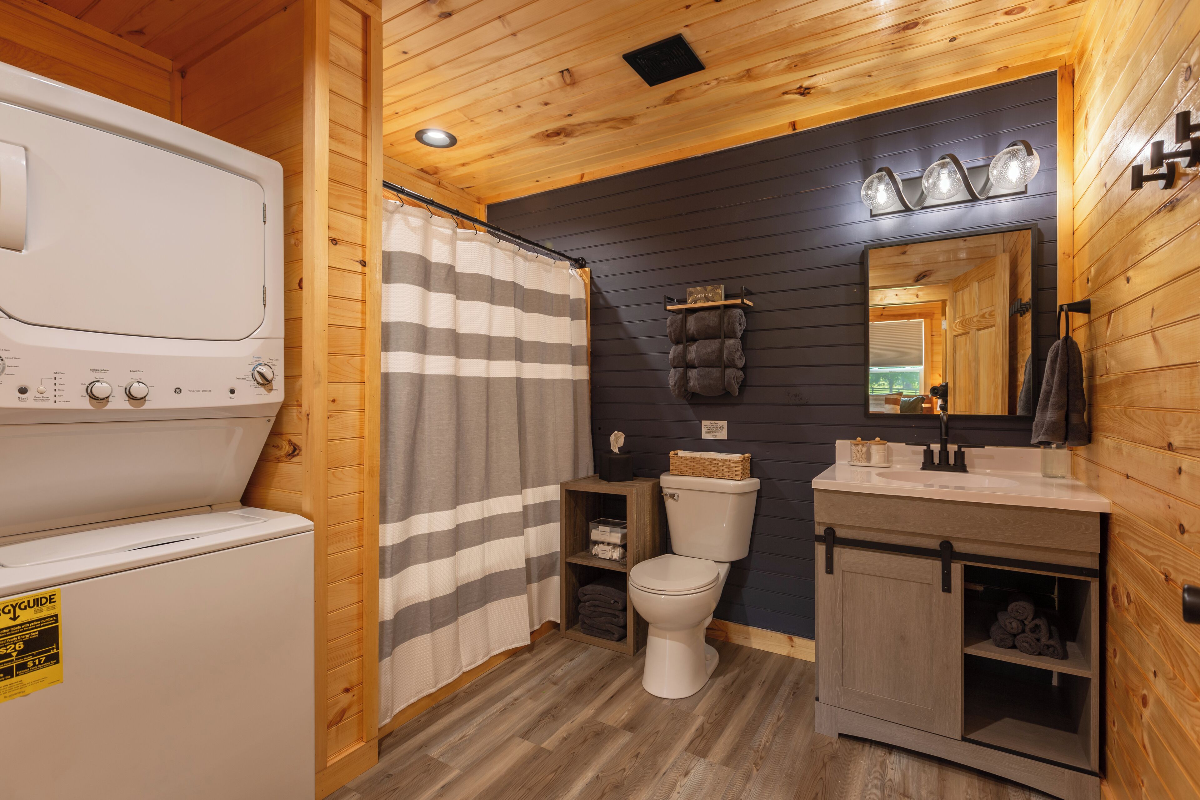 Un baño pequeño con lavadora blanca, cortina de ducha a rayas, inodoro y tocador con espejo.