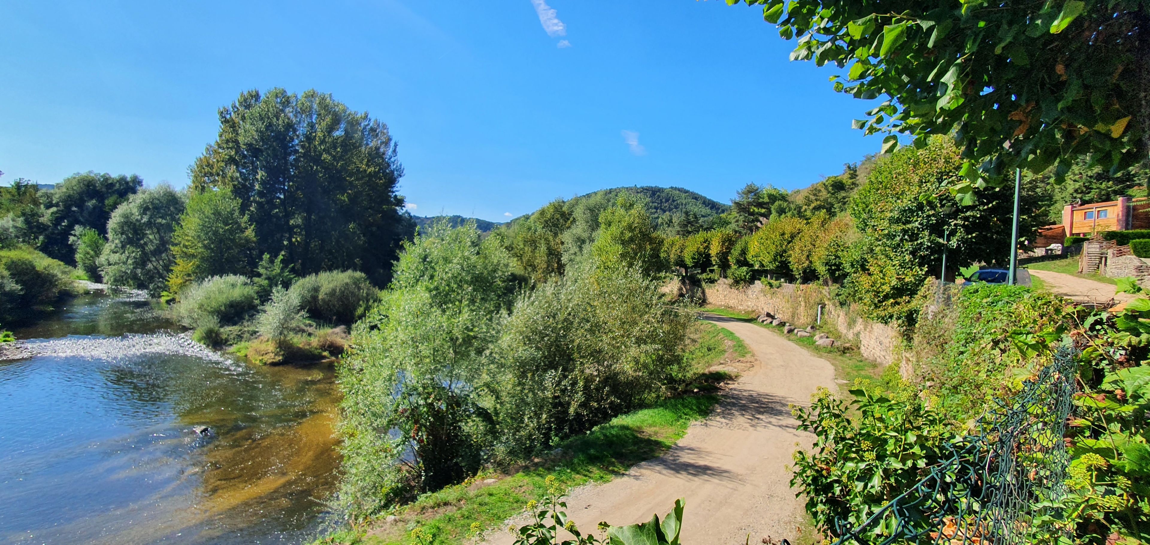 A river with a dirt path alongside it, surrounded by greenery and trees.