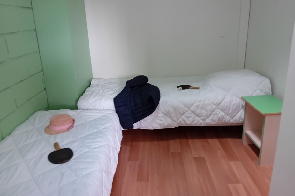A small, compact room with two single beds, a wooden floor, and a green wall.