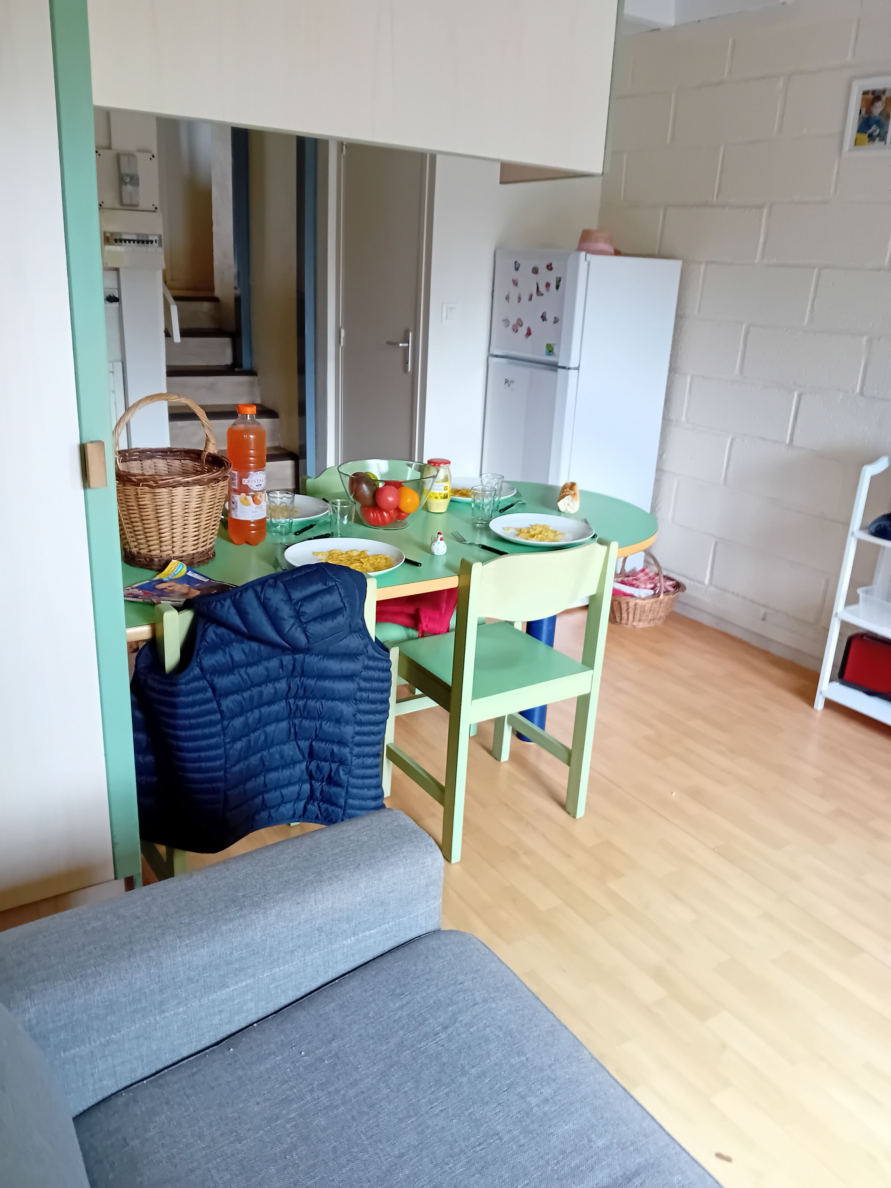 A small, cozy room with a dining table set for two, a green chair, a blue jacket hanging on the chair, and a wooden floor.