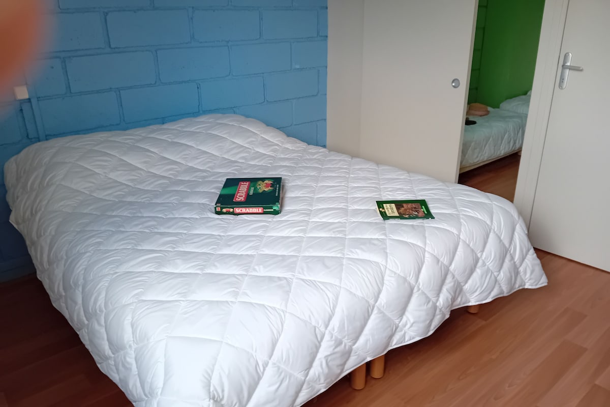 Una habitación con una colcha blanca acolchada, dos cajas del juego Scrabble, piso de madera, pared de ladrillo azul y una puerta verde.