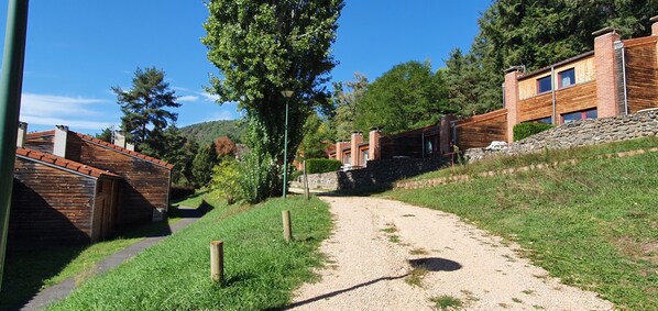 Un chemin de gravier mène à un bâtiment doté d’un mur en pierre et d’un revêtement en bois, le tout étant entouré de verdure et d’arbres.